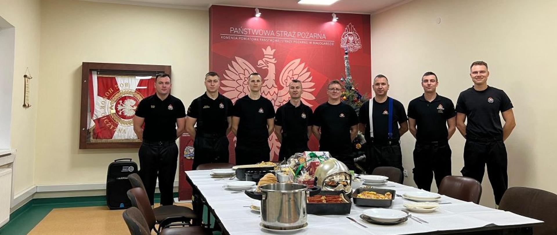Zdjęcie wykonane na świetlicy Komendy. Przy stole wigilijnym 8 strażaków. W tle ścianka z godłem Polski.