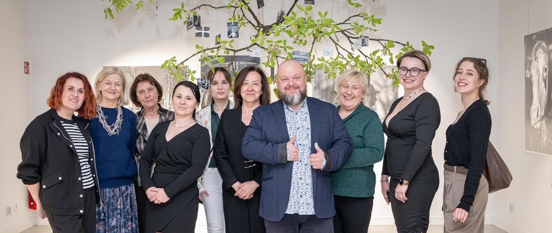 Zdjęcie przedstawia słuchaczki, jednego słuchacza oraz nauczycielki Studium pozujące na tle czarno-białych zdjęć. Nad nimi wisi gałąź drzewa z zielonymi liśćmi.