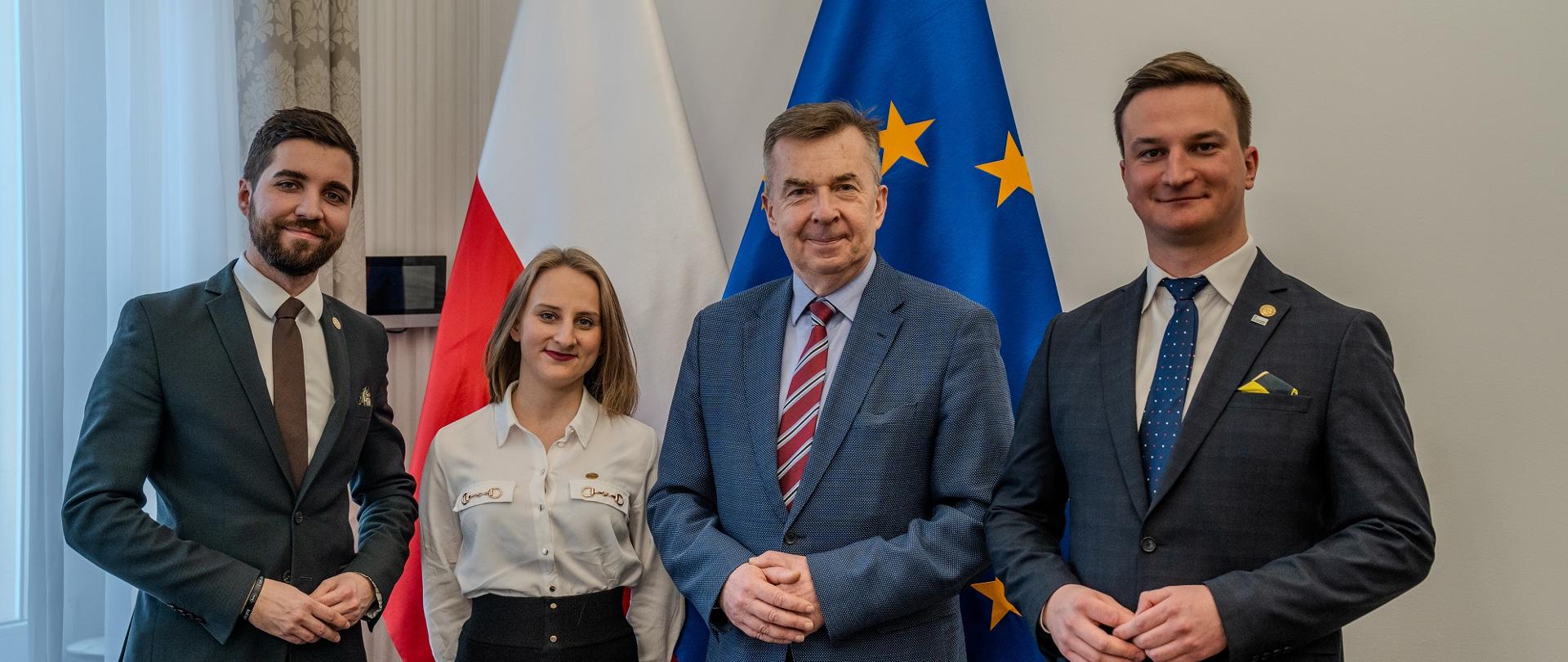 Minister Wieczorek stoi obok dwóch mężczyzn w garniturach i kobiety w białej bluzce, za nimi pod ścianą flagi Polski i UE.