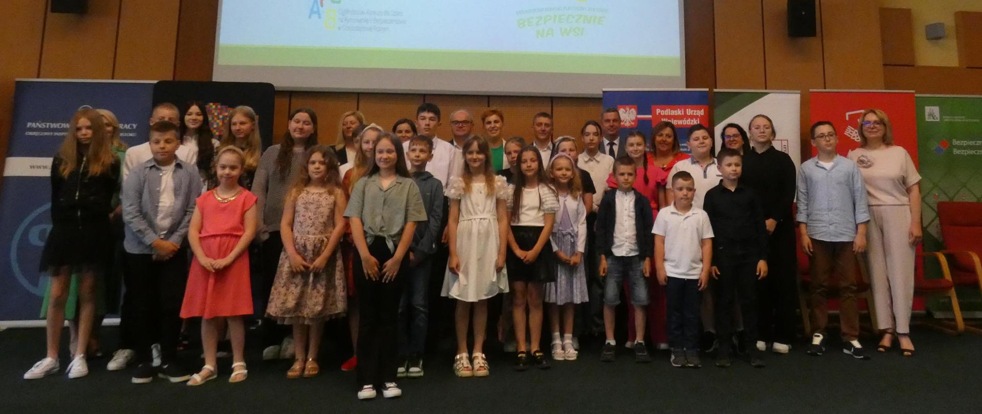 Grupa dzieci pozuje do zdjęcia na tle ekranu z logo KRUS i logo konkursów KRUS dla dzieci. z tyłu przedstawiciel Organizatorów i sponsorów.