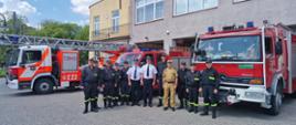 Zdjęcie pamiątkowe uczestników spotkania w OSP w Mszczonowie na tle strażnicy i samochodów gaśniczych przed garażem.