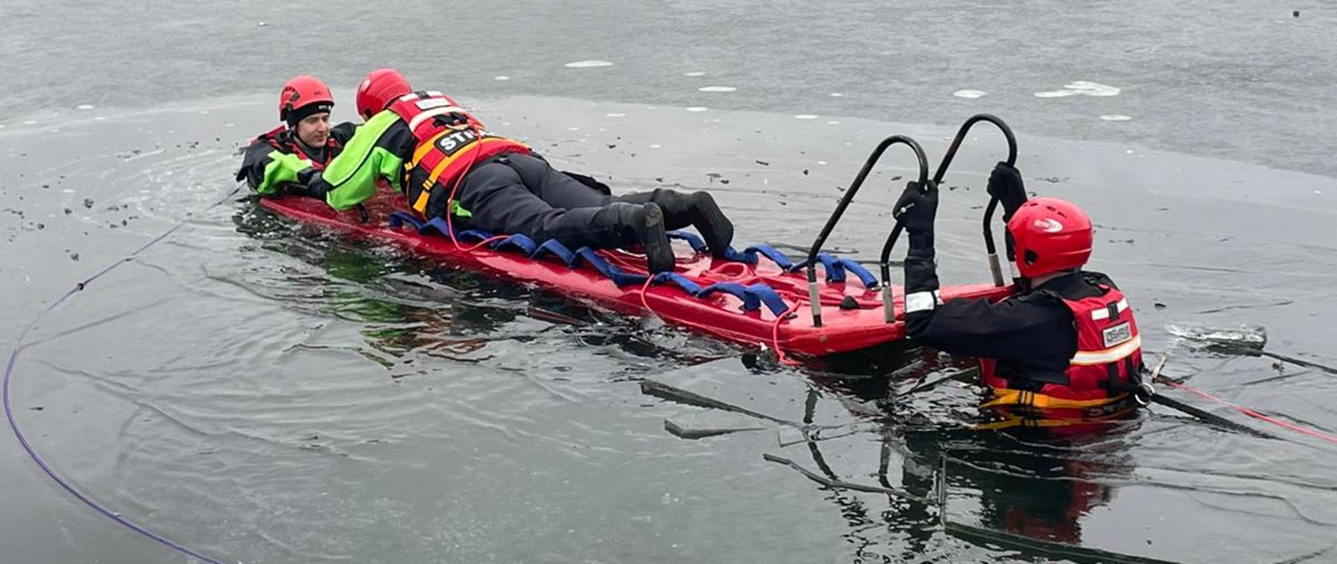 Na zamarzniętym stawie odbywają się pokazy z ratownictwa na lodzie. Dwóch ratowników w kaskach i kombinezonach ochronnych znajduje się na specjalnej desce ratowniczej, częściowo zanurzonej w przerębli. Są przypięci linami i są ciągnięci w stronę brzegu. W tle, na brzegu zbiera się grupa obserwatorów i służb ratunkowych – część osób stoi przy trzcinach, a dalej widać samochody i strażaków. Scena wygląda jak ćwiczenia lub prawdziwa akcja ratunkowa na lodzie.