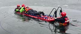 Na zamarzniętym stawie odbywają się pokazy z ratownictwa na lodzie. Dwóch ratowników w kaskach i kombinezonach ochronnych znajduje się na specjalnej desce ratowniczej, częściowo zanurzonej w przerębli. Są przypięci linami i są ciągnięci w stronę brzegu. W tle, na brzegu zbiera się grupa obserwatorów i służb ratunkowych – część osób stoi przy trzcinach, a dalej widać samochody i strażaków. Scena wygląda jak ćwiczenia lub prawdziwa akcja ratunkowa na lodzie.