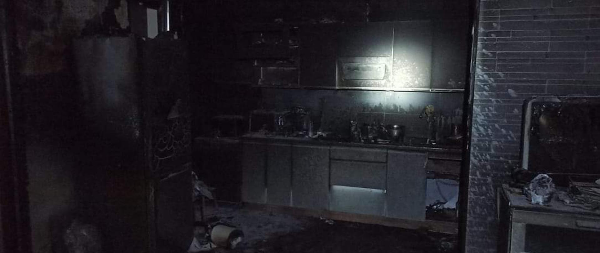 Na zdjęciu znajduje się kuchnia po pożarze. W pomieszczeniu widoczne są meble z frontami w kolorze białym oraz szara lodówka. Po prawej stronie zdjęcia znajduje się komoda wraz z ekranem komputera. Obok komody na podłodze stoi zgrzewka butelek z napojami. Na podłodze są znajdują się porozrzucane przedmioty. Z sufitu oraz z ściany znajdującej się za lodówką po lewej stronie zdjęcia poodpadał tynk. Wszystkie wymienione elementy są pokryte sadzą w kolorze czarnym. W pomieszczeniu panuje ciemność, centralna cześć zdjęcia jest rozświetlona, gdyż została oświetlona przez strażaka przy użyciu latarki. 