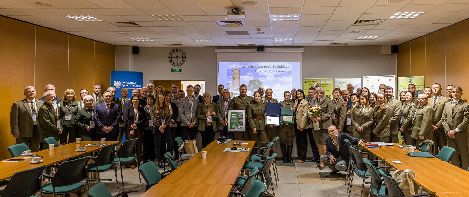 Grupa ludzi, duża część w zielonych mudurach. Patrzą w obiektyw z uśmiechem. Cześć trzyma dyplomy lub kwiaty.
Za nimi wyświetlony slajd z napisem 30-lecie Spalskiego Parku Krajobrazowego