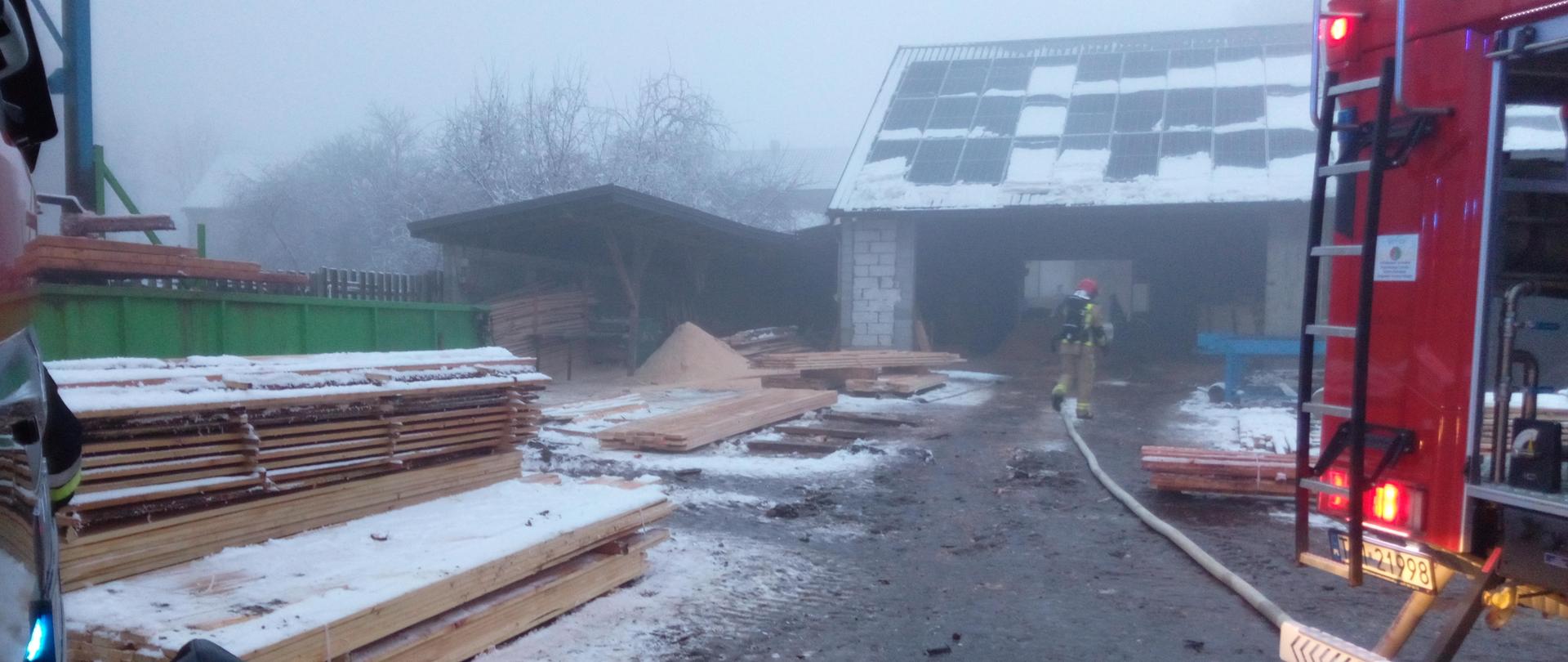 Zdjęcie przedstawia tartak - po lewej ułożone stosy desek, po prawej część samochodu gaśniczego z rozwiniętą linią gaśniczą. W oddali widać strażaka w aparacie ODO. Panuje delikatne zadymienie. nie widać ognia.