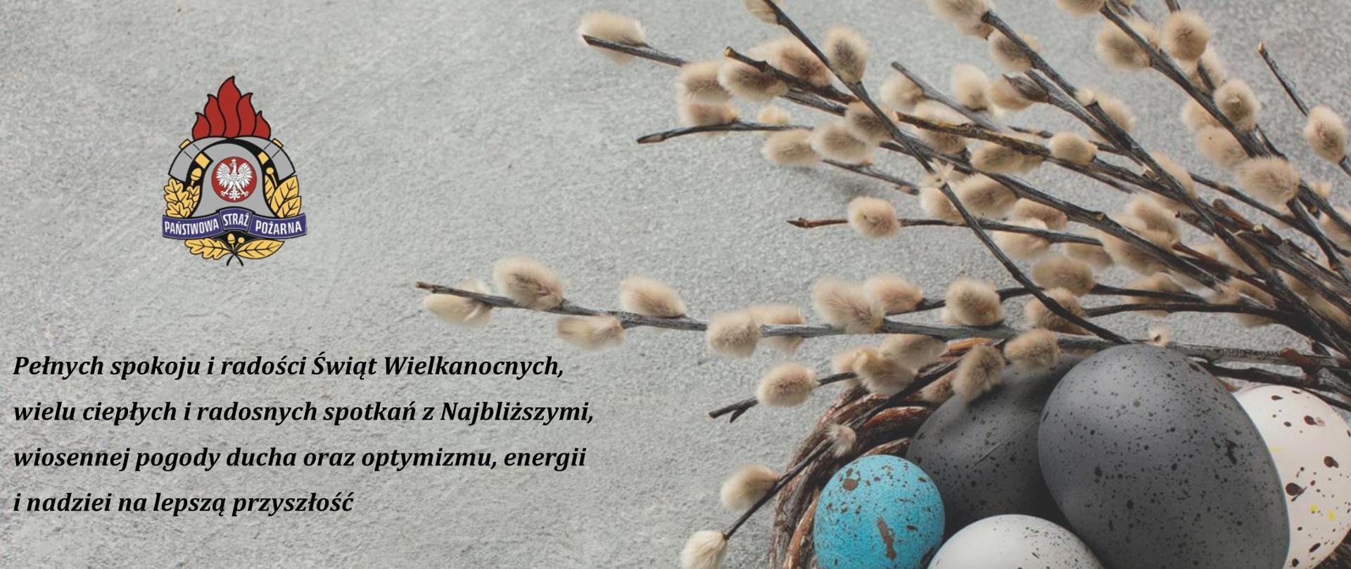 Na szarym tle logo Państwowej straży pożarnej, niżej tekst życzeń świątecznych komendanta powiatowego psp w brzezinach starszego brygadiera mariusza kaczmarskiego. Po prawej stronie zdjęcia koszyk z kolorowymi pisankami świątecznymi