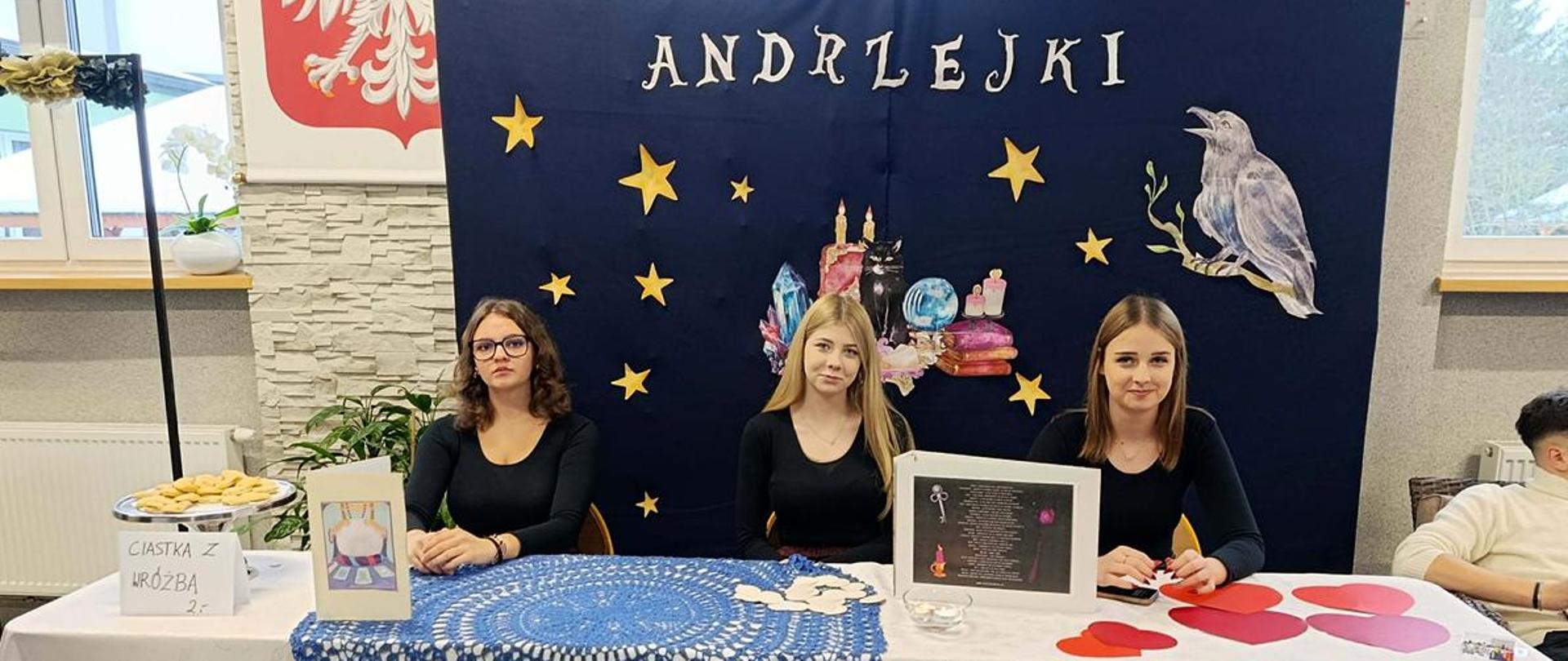 Trzy dziewczyny siedzą przy stole udekorowanym symbolami kojarzącymi się z andrzejkowymi wróżbami, w tle baner z napisem "Andrzejki".