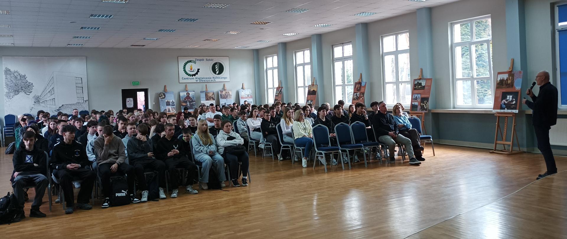 uczniowie Zespołu Szkół Centrum Kształcenia Rolniczego w Oleszycach zgromadzeni w auli szkoły. Przed nimi stoi edukator Instytutu Pamięci Narodowej . Na tylnej ścianie widać logo ZSCKR