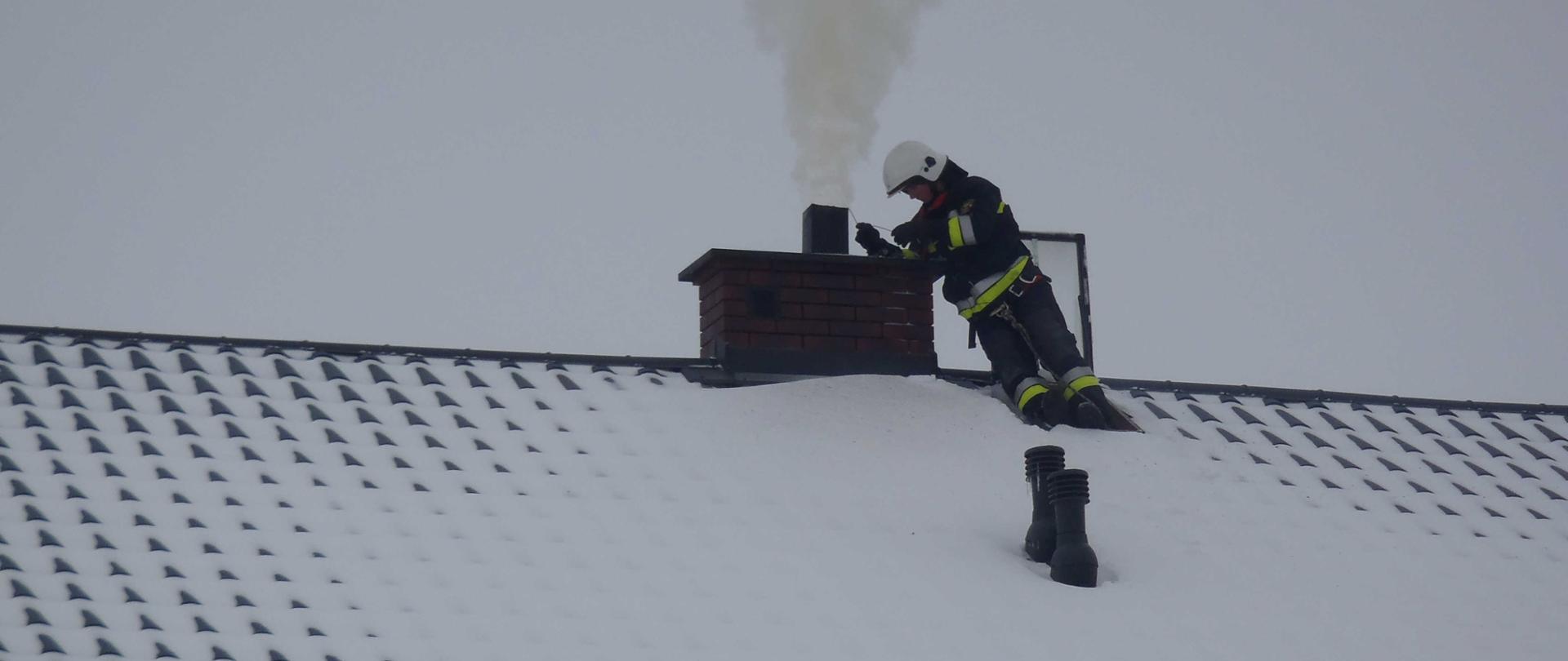 Zdjęcie przedstawia ośnieżony dach domu jednorodzinnego, na którym znajduje się strażak wykonujący czyszczenie komina. 