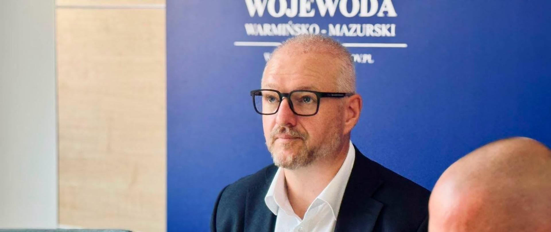 Mężczyzna w okularach, ubrany w granatową marynarkę i białą koszulę, wojewoda, siedzi przed laptopem i obserwuje transmisję wideokonferencji z Ministerstwem Spraw Wewnętrznych i Administracji.