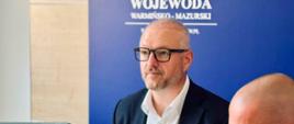 Mężczyzna w okularach, ubrany w granatową marynarkę i białą koszulę, wojewoda, siedzi przed laptopem i obserwuje transmisję wideokonferencji z Ministerstwem Spraw Wewnętrznych i Administracji.