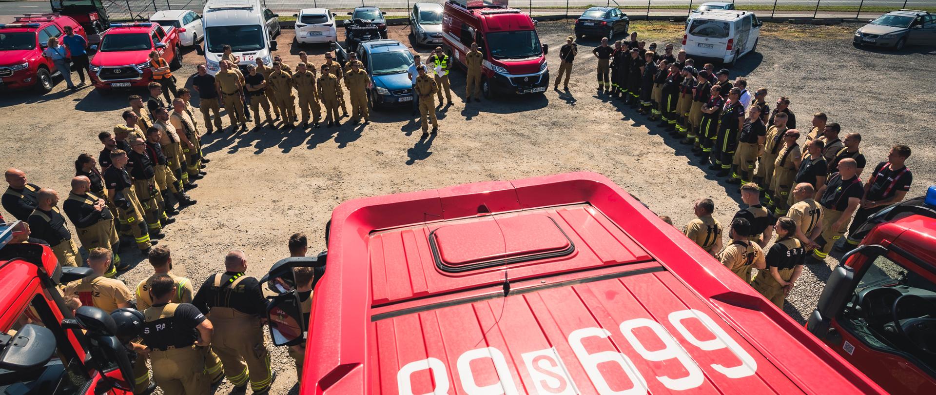 Zdjęcie lotnicze wykonane z perspektywy drona, ukazujące zgrupowanie strażaków na placu manewrowym. Na pierwszym planie widać dach czerwonego wozu bojowego o numerach operacyjnych "89s 699". Strażacy, ubrani w jednolite piaskowe mundury służbowe, stoją w dwóch formacjach półkola wokół centralnego punktu, tworząc rodzaj areny. Pośrodku znajduje się grupa dowódców i instruktorów, w tym dwóch strażaków w czerwonych mundurach bojowych, którzy wydają instrukcje. Za strażakami widać parkujące pojazdy: wóz bojowy, kilka samochodów terenowych i osobowych. W tle rozciąga się pas zieleni i gęsty las pod czystym błękitnym niebem. W prawym dolnym rogu znajduje się logotyp oraz podpis: „st. ogn. Marcin Herok | Państwowa Straż Pożarna”.