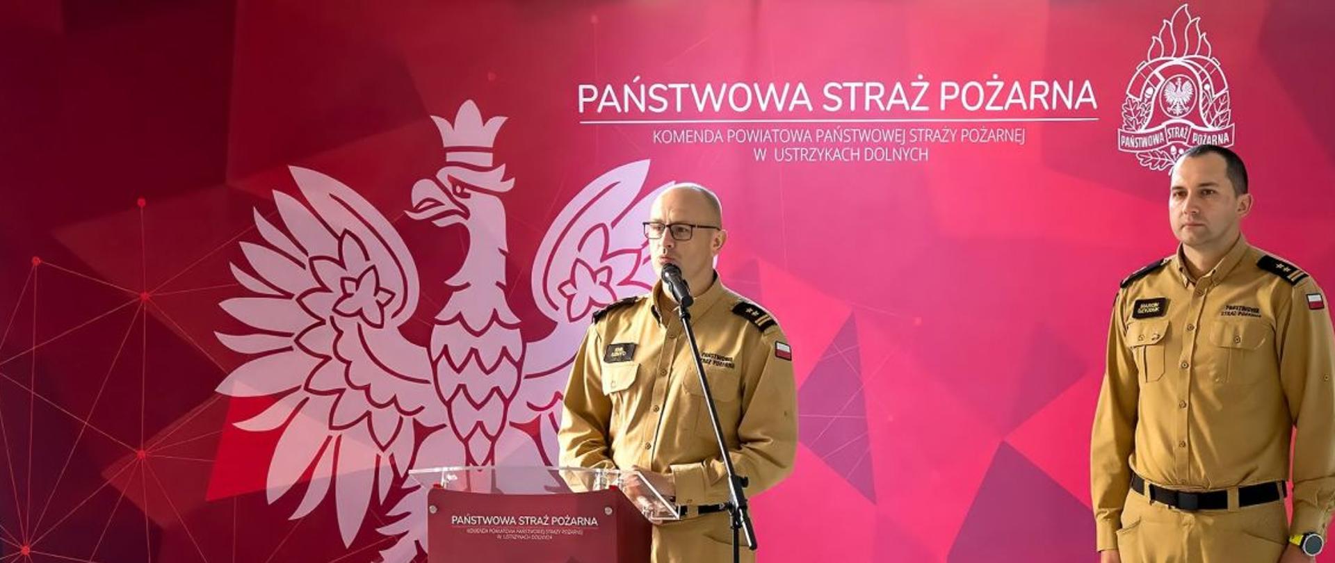 Zdjęcie przedstawia komendanta powiatowego który przemawia przy mównicy, obok stoi zastępca komendanta, w tle logo Państwowej Straży Pożarnej i Orzeł Biały. 