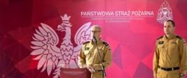 Zdjęcie przedstawia komendanta powiatowego który przemawia przy mównicy, obok stoi zastępca komendanta, w tle logo Państwowej Straży Pożarnej i Orzeł Biały. 