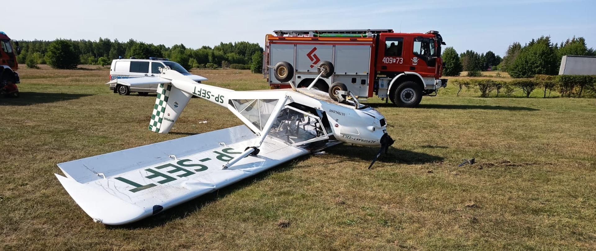 Fotografia przedstawia dwuosobowy samolot ultralekki po "kapotażu", leżący na dachu na trawiastej płycie lądowiska. Płatowiec jest koloru białego. W tle za samolotem widoczny samochód gaśniczy Państwowej Straży Pożarnej oraz radiowóz policyjny.