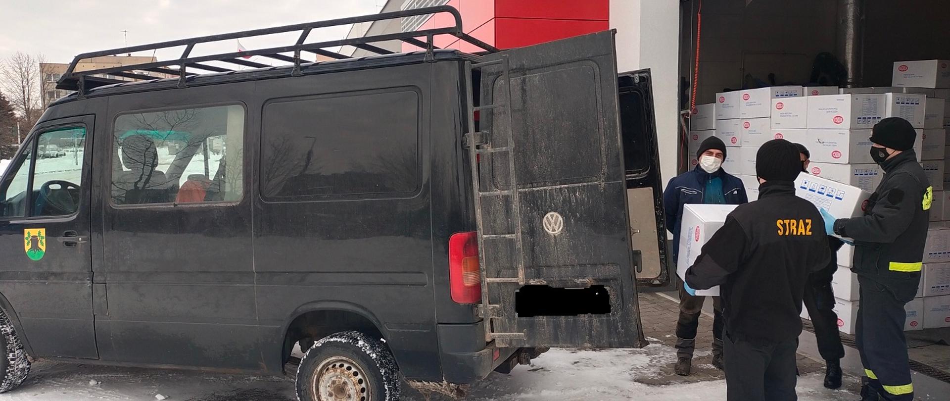 Zdjęcie przedstawia strażaków i inne osoby ładujące kartony z maseczkami do samochodu w tle budynek
