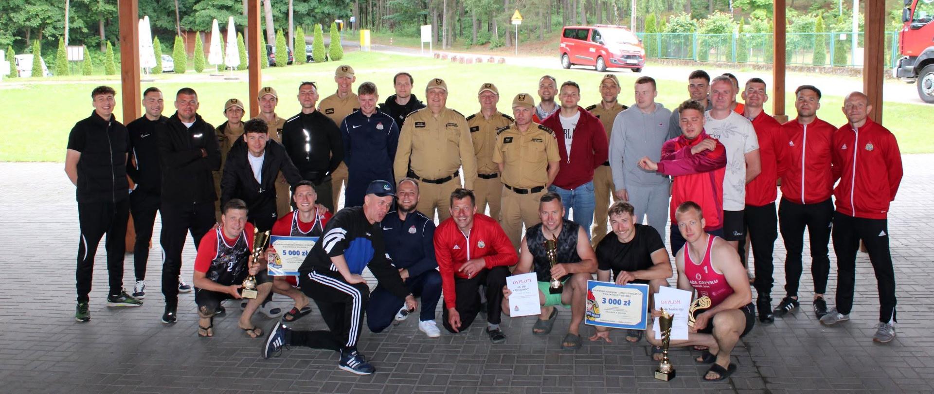 Zdjęcie przedstawia pamiątkową fotografię grupową wszystkich uczestników oraz organizatorów turnieju siatkówki plażowej. Duża grupa mężczyzn pozuje wspólnie pod zadaszoną konstrukcją tarasu, a wśród nich znajdują się zawodnicy w strojach sportowych oraz strażacy w oficjalnych mundurach wyjściowych. Osoby w pierwszym rzędzie klęczą i kucają, dumnie prezentując zdobyte puchar, dyplomy oraz symboliczne czeki z nagrodami finansowymi (widoczne kwoty to 5 000 zł oraz 3 000 zł). W tle, za otwartą przestrzenią wiaty, rozciąga się gęsty zielony las, widać zaparkowane pojazdy, w tym czerwony wóz strażacki, co podkreśla charakter całego wydarzenia.