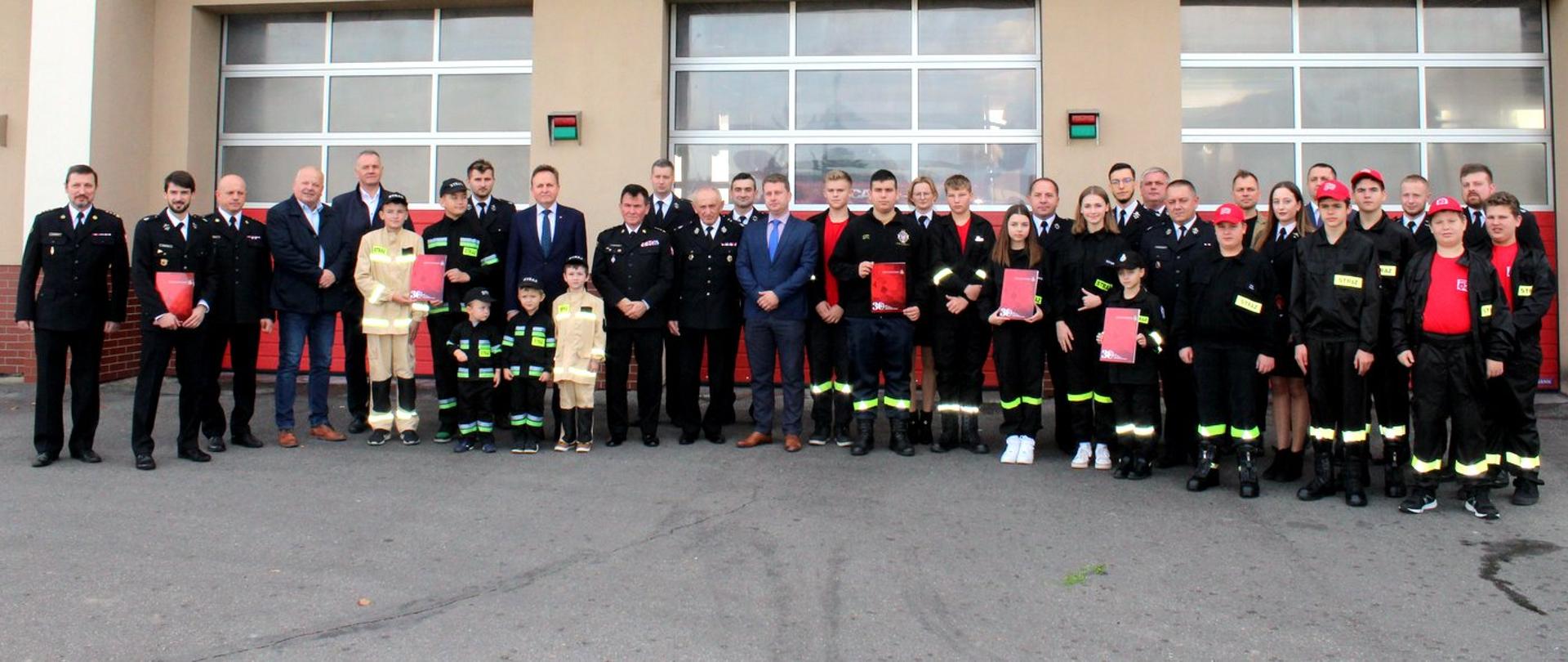Zdjęcie zrobione na zewnątrz przed budynkiem Komendy Powiatowej Państwowej Straży Pożarnej. Na zdjęciu w szeregu stoją uczestnicy uroczystości, wśród nich strażacy ochotniczych straży pożarnych oraz Państwowej Straży Pożarnej. W tle bramy garażowe jednostki ratowniczo-gaśniczej.