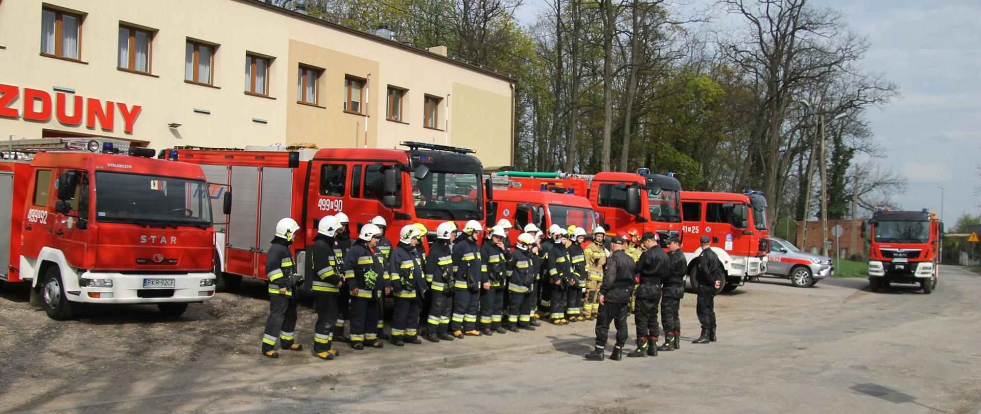 Zdjęcie przedstawia pododdział strażaków stojący przed samochodami bojowymi na tle budynku remizy OSP Zduny, przed strażakami stoją oficerowie Państwowej Straży pożarnej i prowadzą odprawę przed ćwiczeniami.