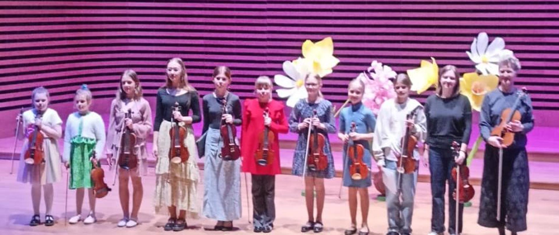 Kobieta i dziesięć uczennic stoją w rzędzie na scenie trzymając w rękach skrzypce.