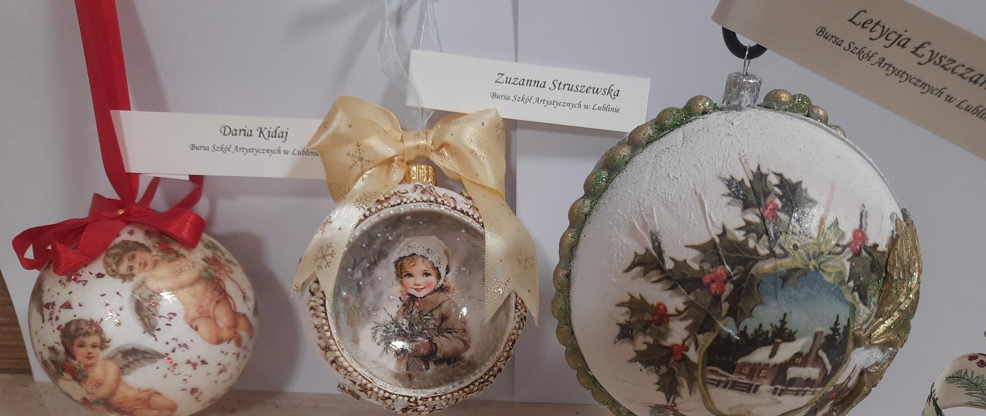 Na jasnym tle są wyeksponowane trzy bombki. Z lewej strony jest biała bombka z aniołkami i czerwoną kokardą podpisana: Daria Kidaj Bursa Szkół Artystycznych w Lublinie. W środku bombka z wizerunkiem małej dziewczynki w zimowym otoczeniu, z kremową kokardą, podpisana: Zuzanna Struszewska Bursa Szkół Artystycznych w Lublinie. Z prawej strony jest biała bombka z obrazkiem zimowej chatki otoczonym gałązkami ostrokrzewu i podpisana: Letycja Łyszczarz Bursa Szkół Artystycznych w Lublinie