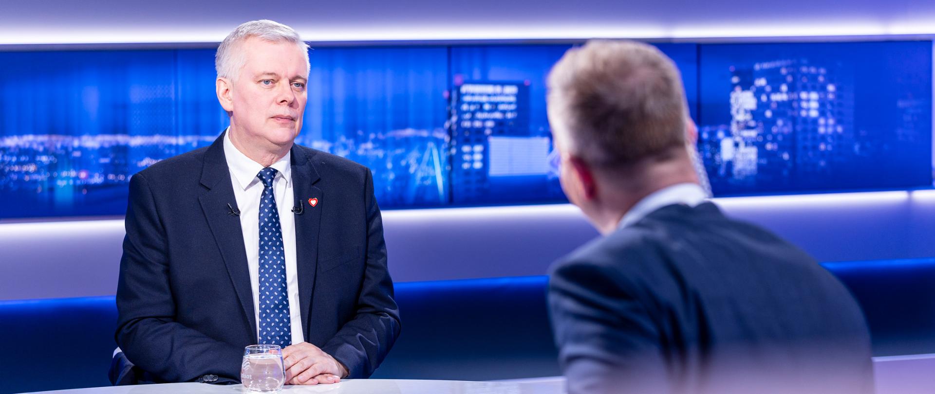 Zdjęcie przedstawia studio telewizyjne. Przy białym stole siedzą dwie osoby - po lewej minister Tomasz Siemoniak, a po prawej redaktor Marek Tejchman (siedzący tyłem). Na stole przed ministrem stoi szklanka z wodą. W tle widać ekrany z panoramą nocnego miasta z oświetlonymi budynkami. 