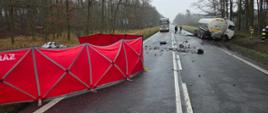 Wypadek na drodze; cysterna stoi w rowie, na jezdni leżą odłamki, a po lewej ustawiono czerwony parawan z napisem straż
