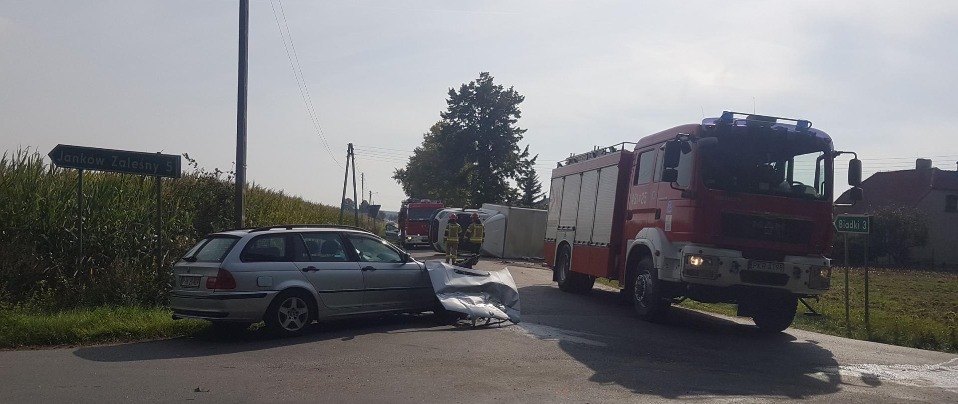 Skrzyżowanie dróg. Tuż przy skrzyżowaniu zaparkowany pojazd pożarniczy. Obok pojazdu uszkodzony samochód osobowy. Pojazd ma uszkodzony przód. Obok BMW leży maska przednia. W oddali widoczny samochód dostawczy leżący na boku oraz dwóch strażaków w ubraniach specjalnych. 