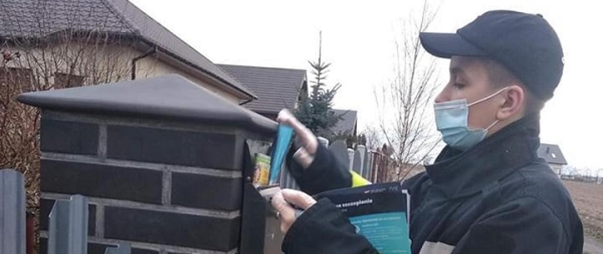 Zdjęcie przedstawia strażaka umieszczającego ulotkę dotyczącą szczepień przeciwko
COVID-19 w skrzynce pocztowej
