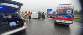 Na zdjęciu strażacy prowadzący działania na autostradzie A1. Obok radiowóz z otwartym bagażnikiem, uszkodzony bus na boku oraz 2 karetki.