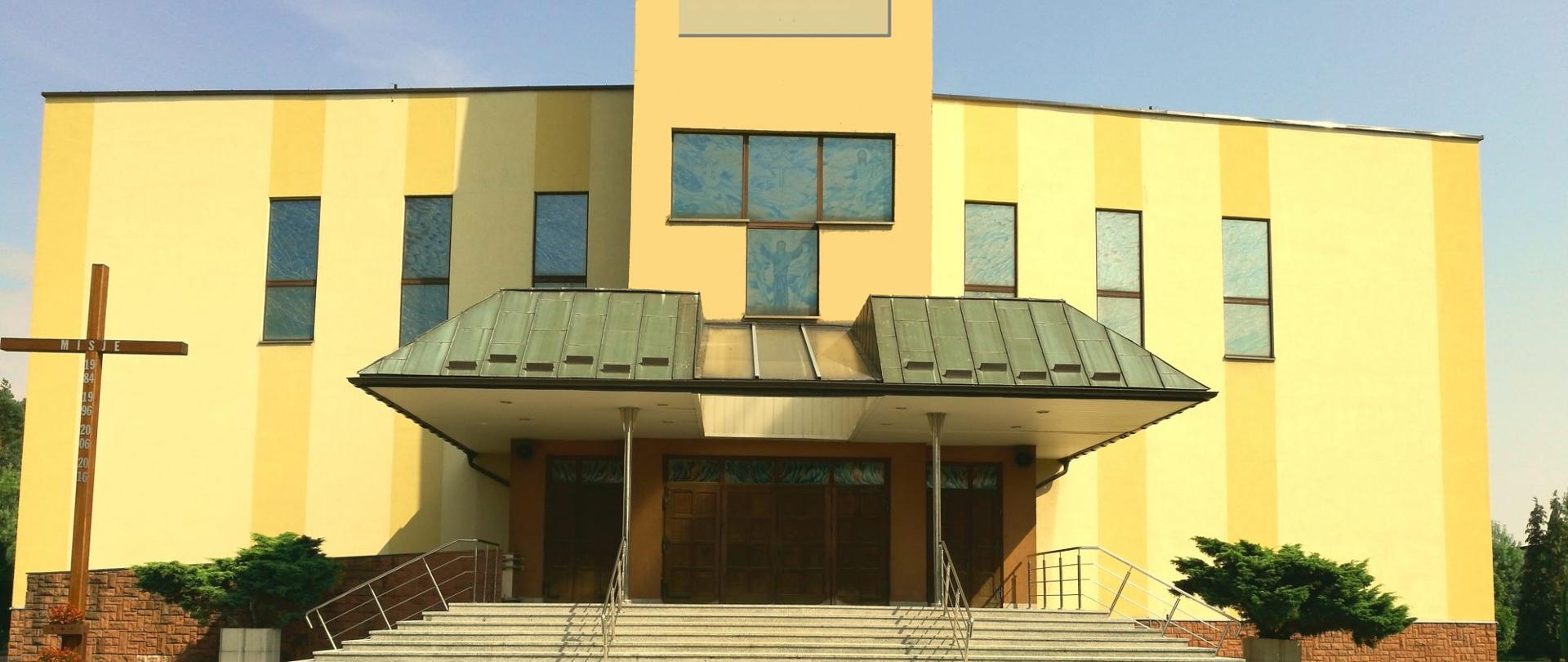Na zdjęciu widać jasny, żółto-kremowy budynek kościoła o prostej, nowoczesnej bryle. Na środku znajduje się podwyższone wejście z szerokimi schodami, metalowymi poręczami i dużym zadaszeniem o ciemnym, spadzistym dachu. Nad wejściem umieszczono wysoką część fasady z oknami. Po obu stronach widoczne są wąskie, pionowe okna.
Przed budynkiem rozciąga się wybrukowany plac. Po lewej stronie stoi duży drewniany krzyż, a przy elewacji znajdują się niskie, przycięte krzewy. W tle widać błękitne niebo i fragmenty zieleni.