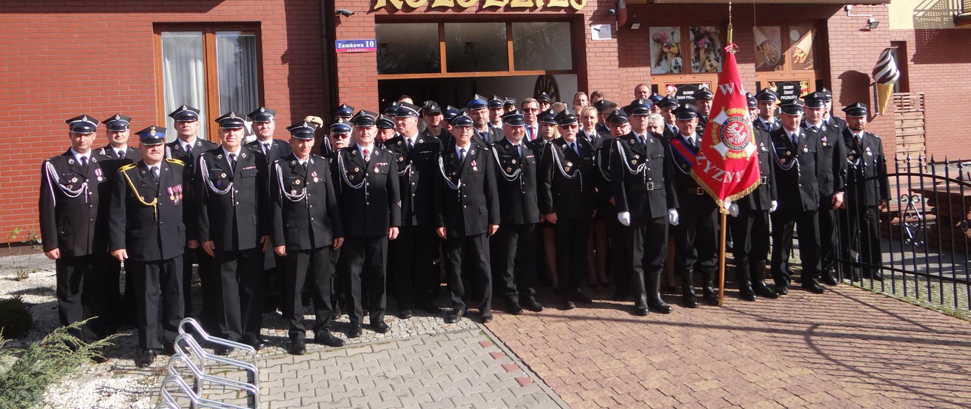 Na zdjęciu przed budynkiem stoją wraz ze sztandarem druhowie OSP oraz strażak PSP w umundurowaniu wyjściowym.