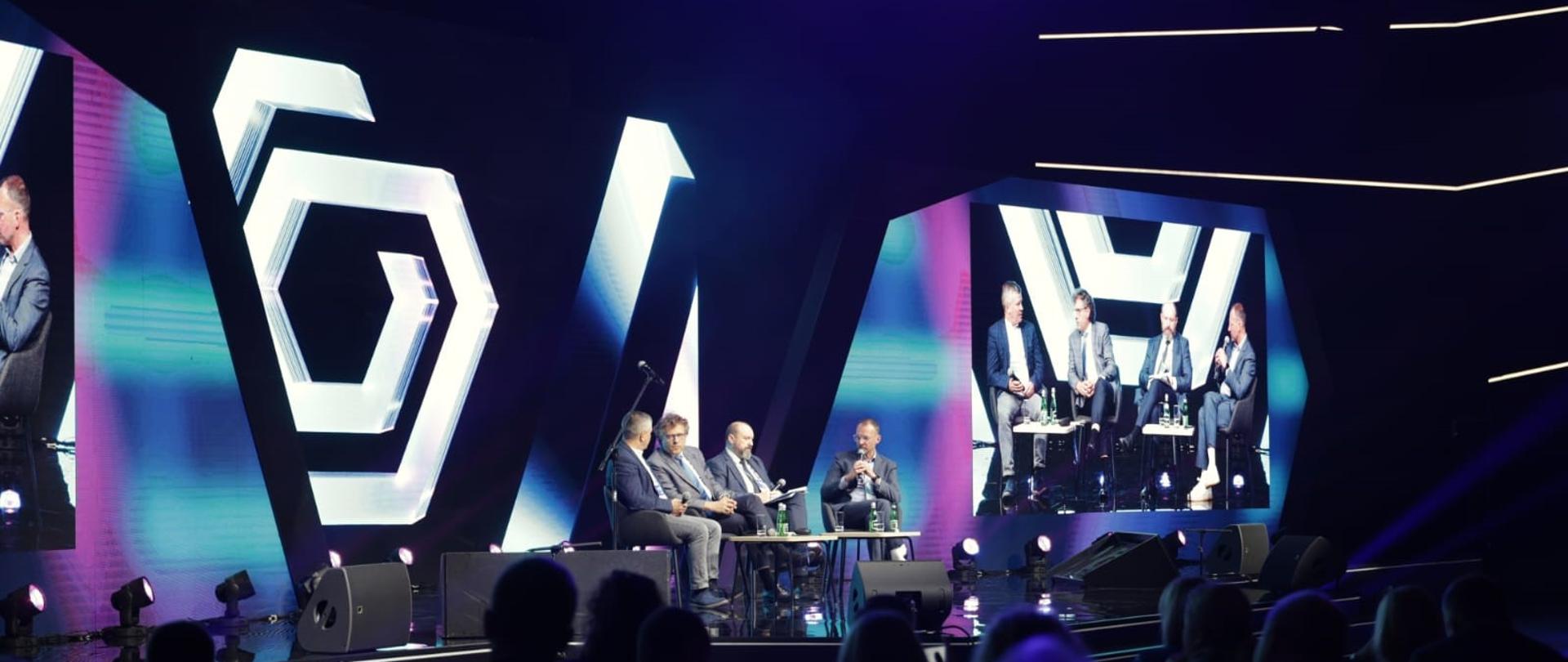 Kilka osób siedzi na scenie na sali. Wśród nich wiceminister Jacek Karnowski.