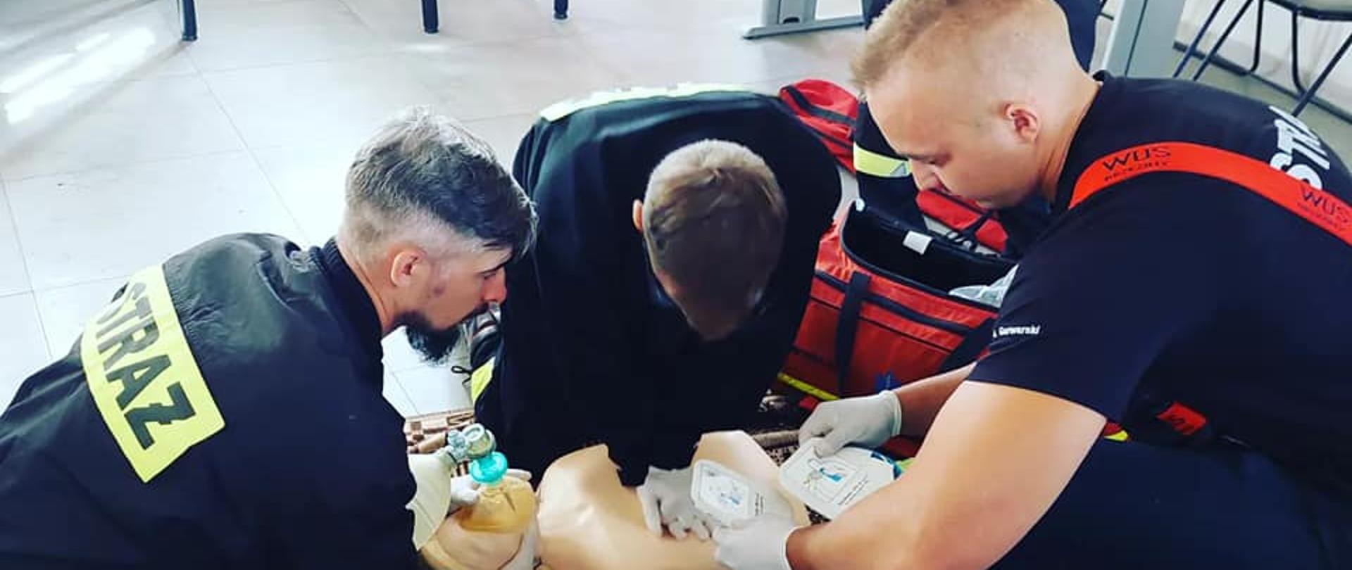 Trzech strażaków udziela pierwszej pomocy manekinowi dorosłej osoby, poprzez wykonanie resuscytacji krążeniowo oddechowej. Jeden ze strażaków trzyma w ręku elektrody od defibrylatora.