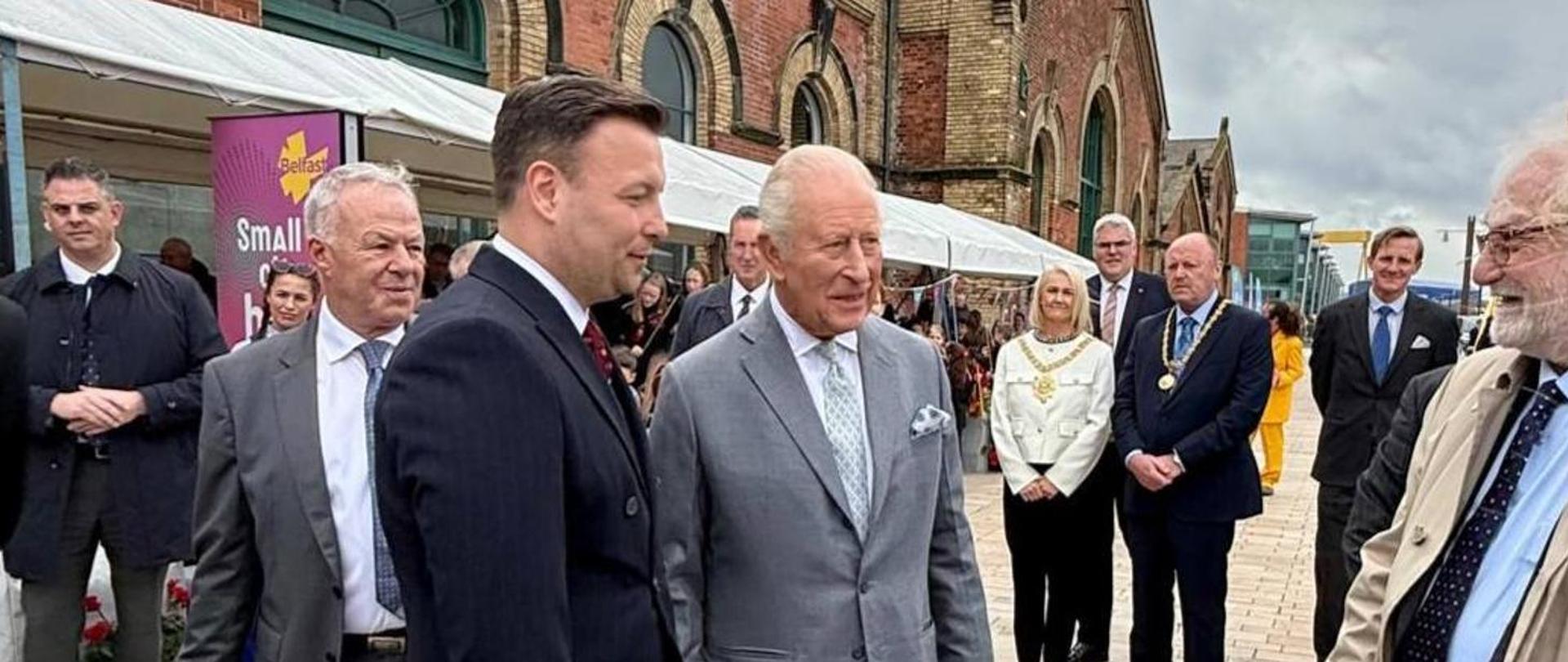 Król Wielkiej Brytanii w jasnoszarym garniturze rozmawia z Konsulem Generalnym RP w Belfaście, Panem Grzegorzem Salą, podczas oficjalnego wydarzenia plenerowego przed zabytkowym ceglanym budynkiem. W tle stoją uczestnicy spotkania, namioty oraz baner z napisem „Belfast”.
