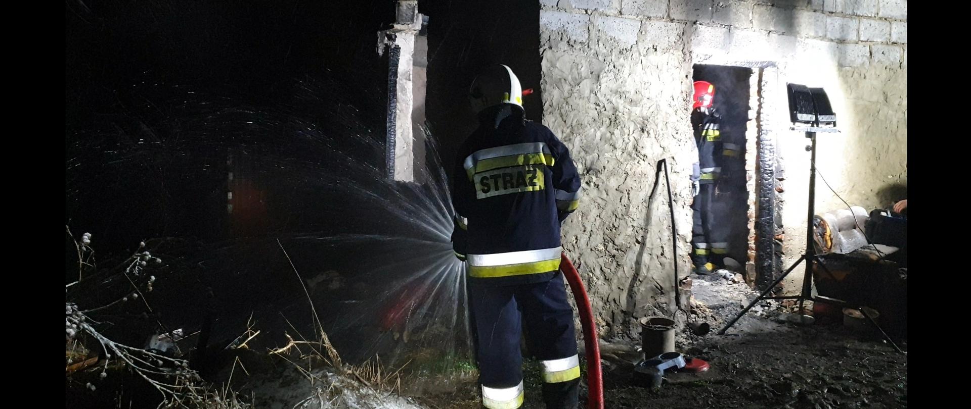 Najaśnica oświetla miejsce pożaru, strażak ubrany w ubranie bojowe przelewa zgliszcza wodą. W tle budynek statyw z najaśnicą. Pora nocna.