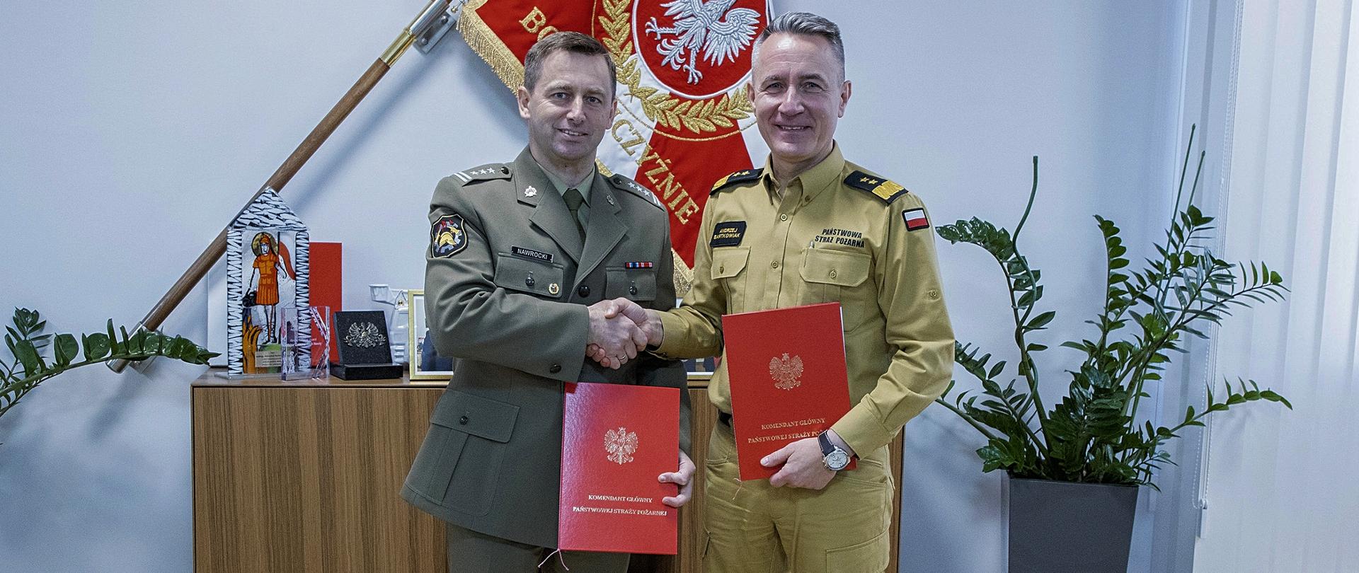 Zdjęcie pamiątkowe. Dwaj mężczyźni w polskich mundurach, trzymający w rękach teczki z porozumieniem, ściska sobie dłonie na tle sztandaru Komendy Głównej Państwowej Straży Pożarnej.