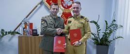 Zdjęcie pamiątkowe. Dwaj mężczyźni w polskich mundurach, trzymający w rękach teczki z porozumieniem, ściska sobie dłonie na tle sztandaru Komendy Głównej Państwowej Straży Pożarnej.