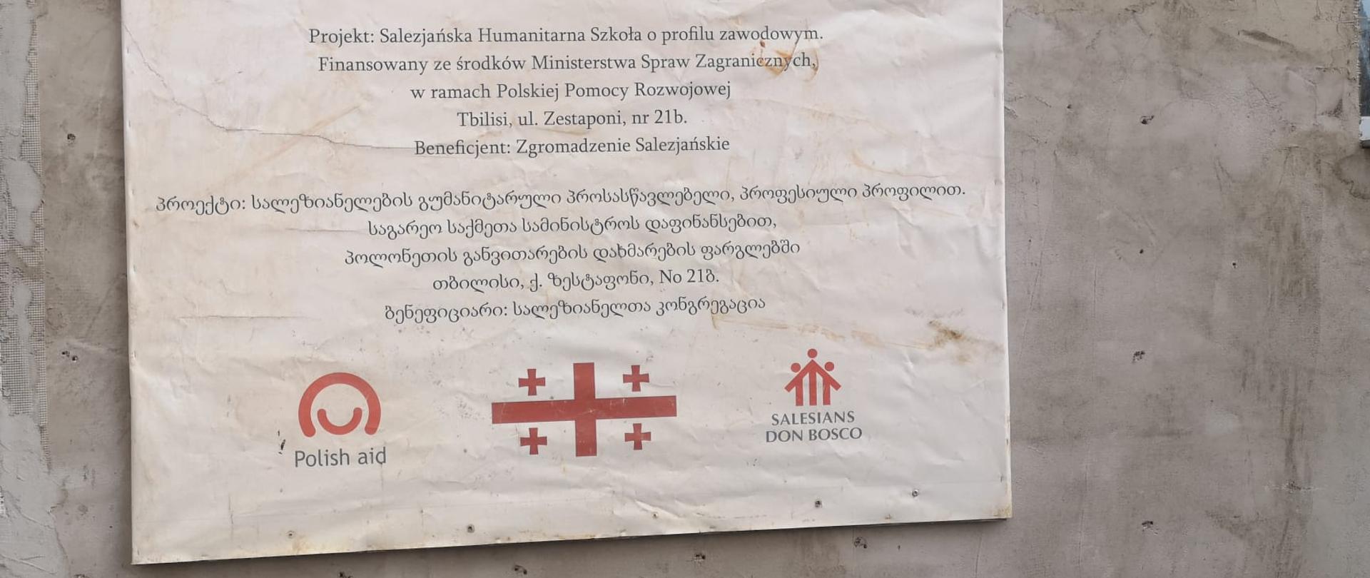 Tablica informacyjna na temat projektu, na której napisane jest: Projekt: Salezjańska Szkoła Humanitarna o profilu Zawodowym. Finansowany ze środków Ministerstwa Spraw Zagranicznych w ramach Polskiej Pomocy Rozwojowej. Napisy po polsku i po gruzińsku. Na dole logo Polskiej pomocy, flaga Gruzji i logo Salezian Don Bosco. 