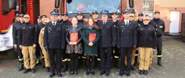 Zdjęcie przedstawia strażaków i pracowników cywilnych podczas uroczystej zmiany służby.
W tle budynki.
