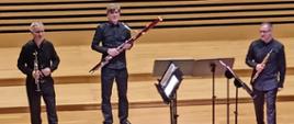 Trzech mężczyzn stoi na estradzie trzymając w rękach instrumenty (klarnet, fagot, obój), zwróceni przodem do publiczności, przed nimi stoją pulpity. 
