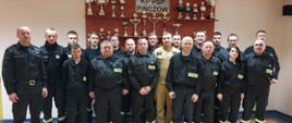 Strażacy ratownicy Ochotniczych Straży Pożarnych z komisją egzaminacyjną na tle gabloty z pucharami KP PSP w Pińczowie