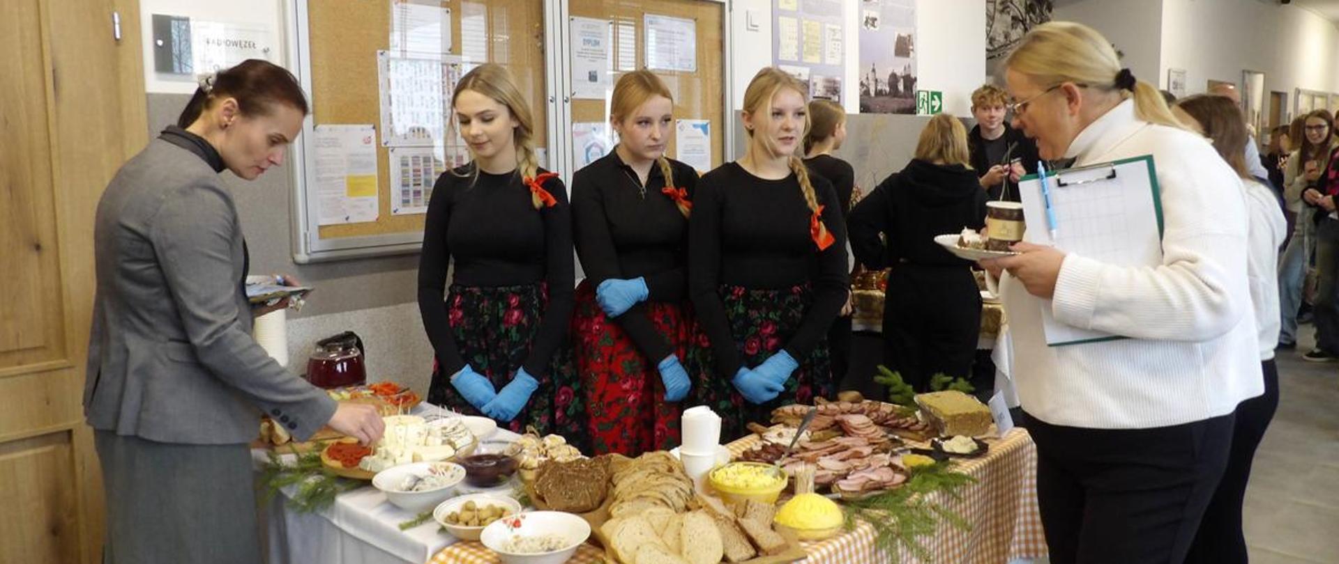 Stoisko gastronomiczne ustawione na korytarzu szkolnym podczas wydarzenia kulturalnego. Trzy uczennice w ludowych spódnicach i czarnych bluzkach prezentują tradycyjne potrawy, w tym wędliny i pieczywo, członkom jury.