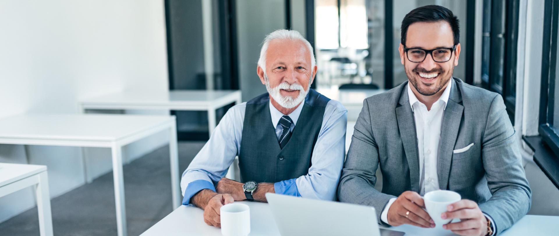 Na zdjęciu widać starszego pana z młodszym panem. Siedzą w biurze przy biurku, przed nimi laptop. Trzymają w ręku kubek.