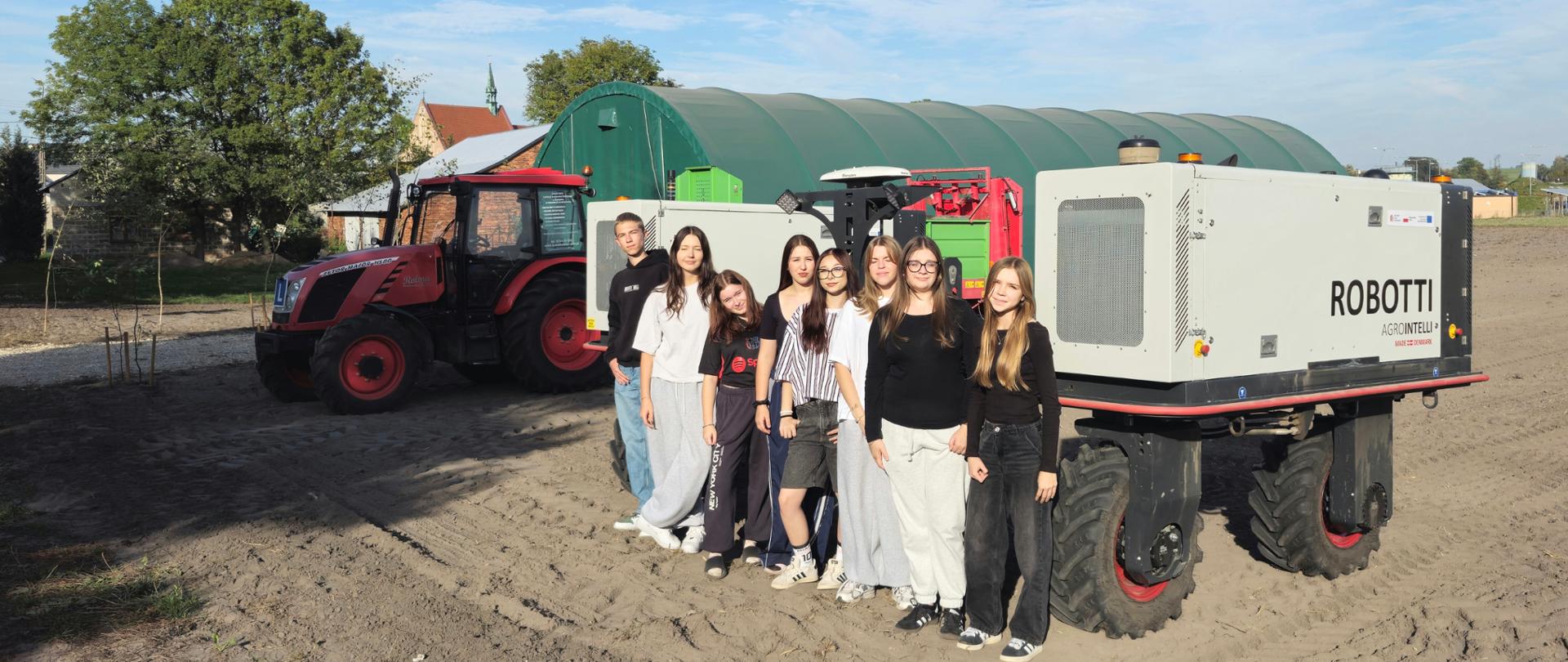 Grupa młodzieży pozuje do zdjęcia na tle ciągnika rolniczego i autonomicznego robota polowego na terenie szkolnego gospodarstwa rolnego.