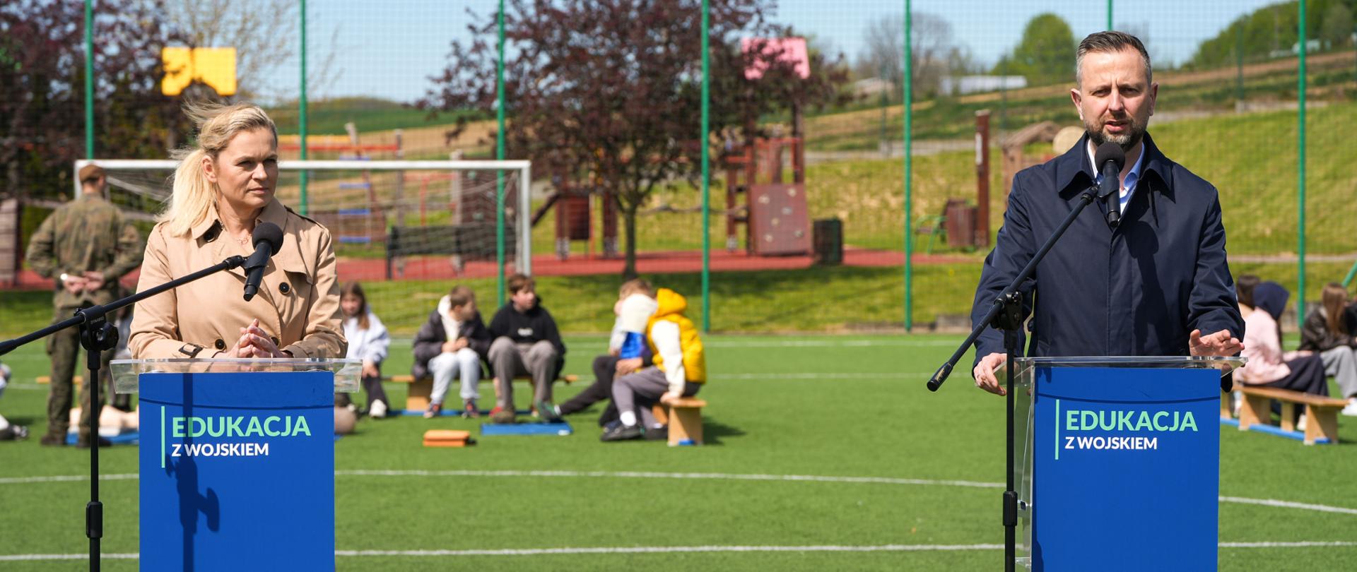 Dwie osoby przemawiają przy niebieskich mównicach z napisem „EDUKACJA Z WOJSKIEM”. Po lewej stronie stoi kobieta w beżowym płaszczu, po prawej mężczyzna w granatowej kurtce. W tle, na trawiastym boisku, widać siedzące na ławkach dzieci oraz żołnierza.