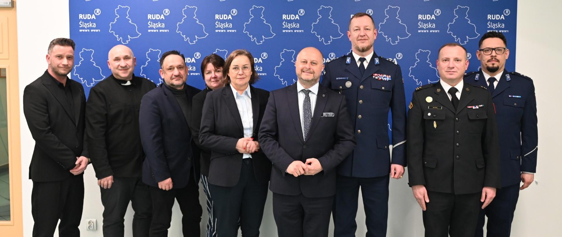 Grupa kobiet i mężczyzn, w tym przedstawiciele samorządu, duchowieństwa oraz służb mundurowych, pozuje do wspólnego zdjęcia na tle ścianki z logo Ruda Śląska w Urzędzie Miasta.