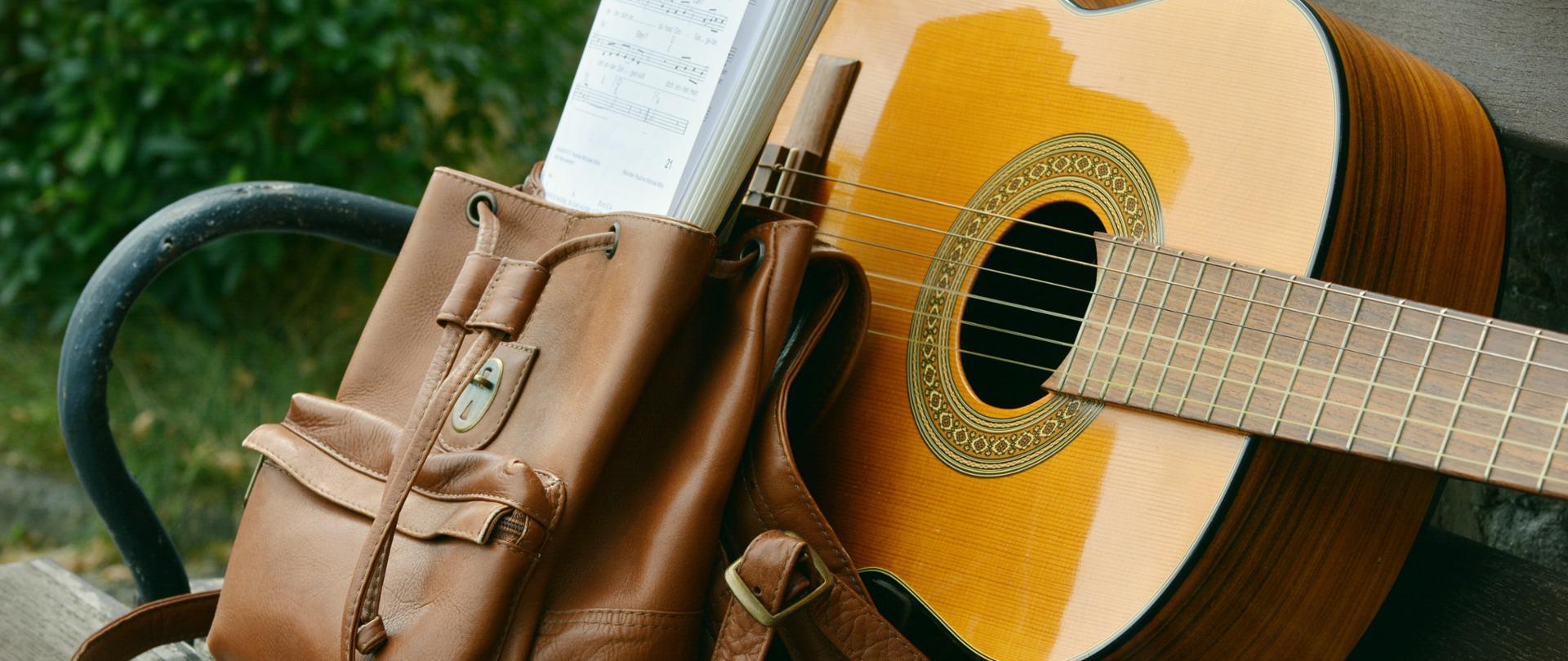 Zdjęcie przedstawiające gitarę leżącą obok plecaka z nutami na ławce.