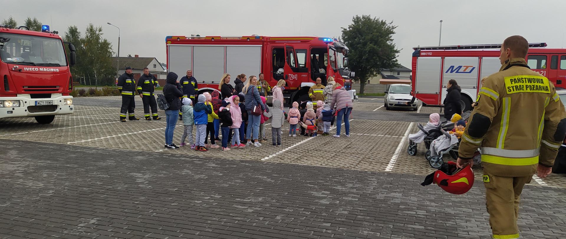 W centralnym punkcie zdjęcia stoją panie wychowawczynie z grupą małych dzieci. Z prawej strony idzie w ich kierunku strażak. W tle stoją trzy bojowe wozy strażackie
